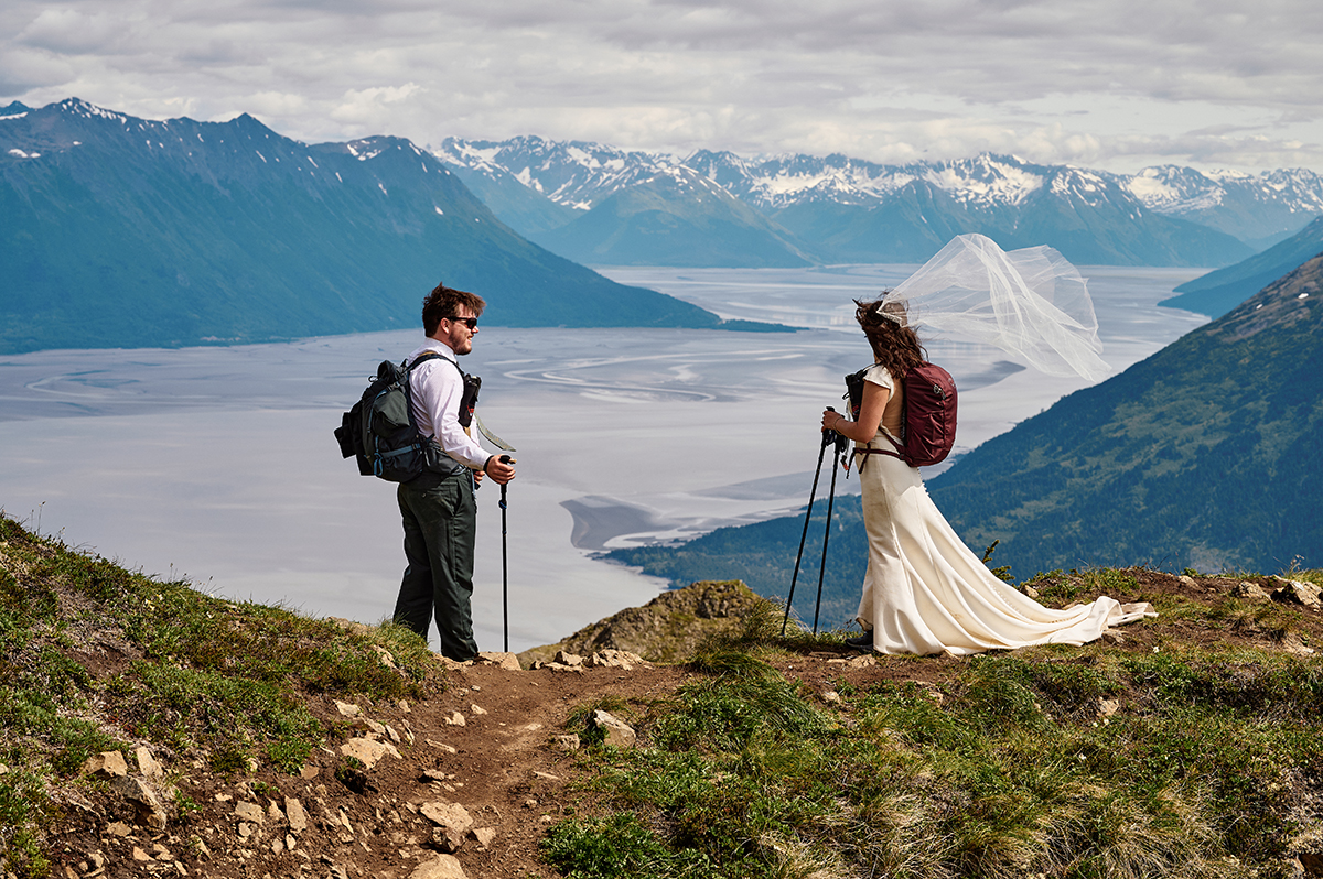 A couple stand atop a mountain overlooking a bay. Both carry backpacks and the bride's veil blows in the wind behind her.